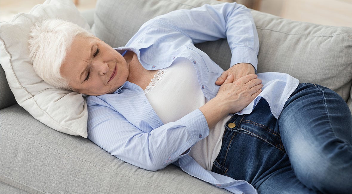 Concerned elderly woman laying on her couch, holding her abdomen, suffering from stomach ache