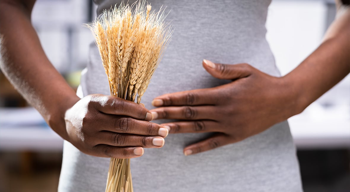 woman in a gray shirt holding her stomach in one hand and a spikelet of wheat in the other.