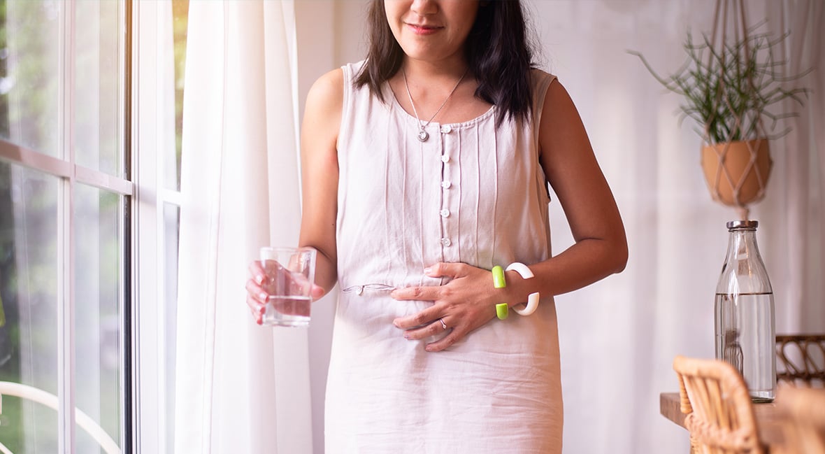 woman in white dress holding a glass of water in one hand and clutching her stomach in pain with the other